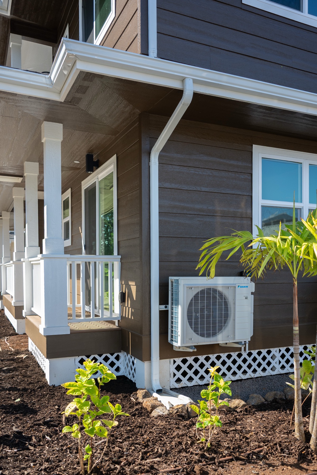 Mini split condenser unit installed on the side of a Hawaii home, with a covered porch and palm trees